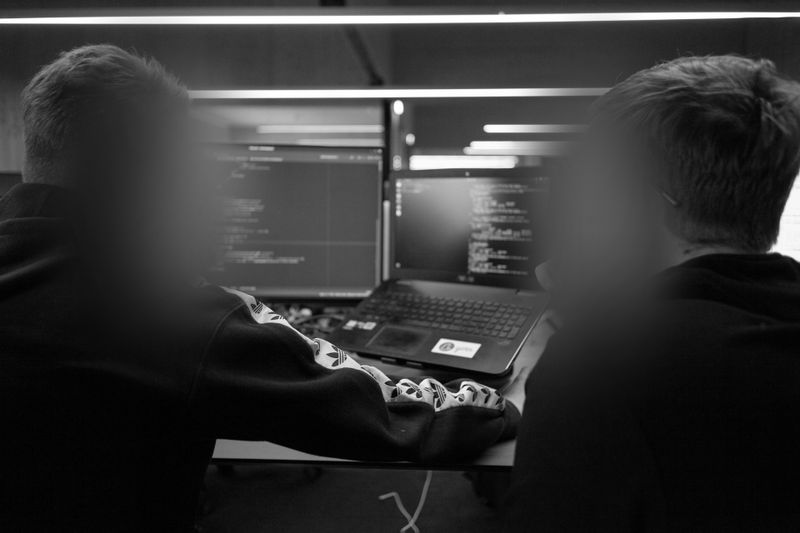Zwei Personen mit verschwommenen Gesichtern arbeiten an einem Schreibtisch mit mehreren Computermonitoren an Programmierung in einem schwach beleuchteten Raum.