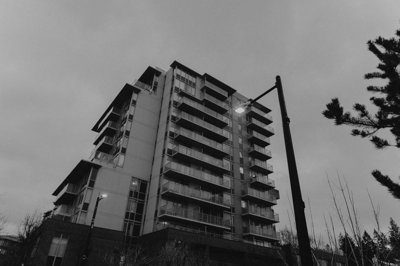 Photo en noir et blanc d’un immeuble résidentiel de plusieurs étages sous un ciel nuageux, avec un lampadaire allumé au premier plan.