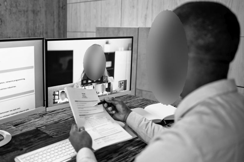 foto en blanco y negro de un empleado de oficina de piel oscura sosteniendo un montón de hojas de papel en la mano, leyendo y siguiendo con el bolígrafo, durante una videollamada, las caras en esta foto han sido anonimizadas