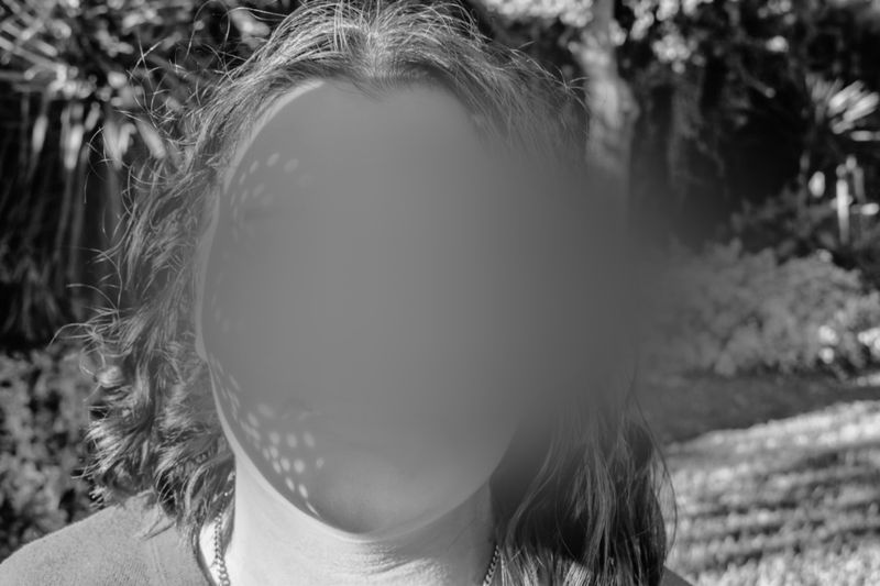 A person with long hair standing outdoors, face blurred, surrounded by trees and sunlight creating dappled shadows. Black and white image.