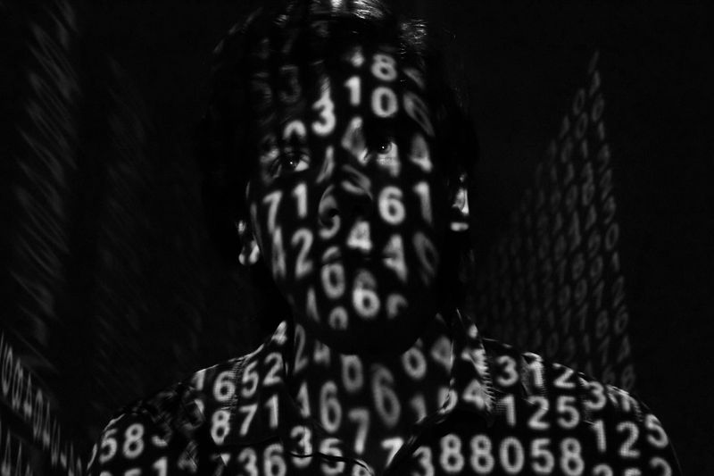 Le visage d'une personne est masqué par des chiffres projetés, créant un effet numérique et mystérieux dans un décor sombre.