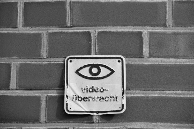 Czarno-biały znak z symbolem oka i napisem „video-überwacht” na ceglanej ścianie, wskazujący na monitoring wideo.
