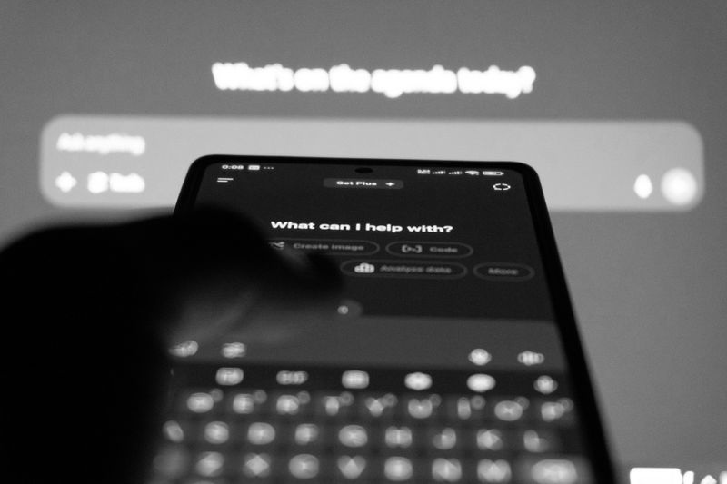 Foto en blanco y negro, en la que aparece un teléfono con una aplicación de chat IA abierta, tocando con el dedo, sobre el fondo de la pantalla con la misma aplicación
