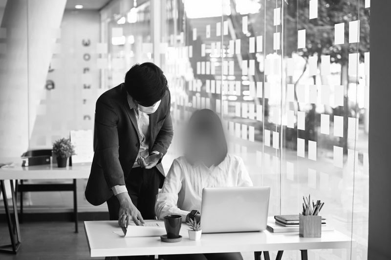 Deux personnes dans un bureau : l'une debout, pointant un document, l'autre assise à un bureau avec un ordinateur portable. Image en noir et blanc.