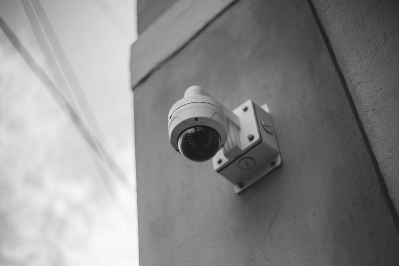 Image en noir et blanc d'une caméra de sécurité montée sur un coin de bâtiment, légèrement inclinée vers le bas.