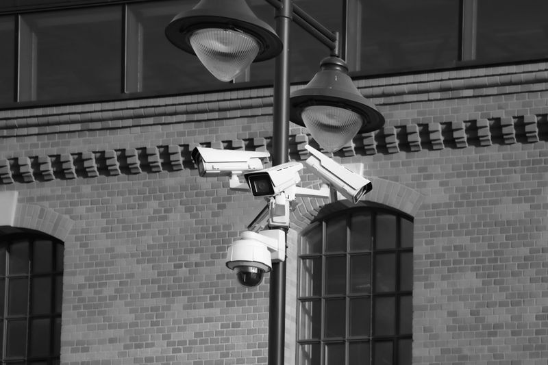Image en noir et blanc de plusieurs caméras de sécurité montées sur un poteau devant un bâtiment en briques avec de grandes fenêtres.