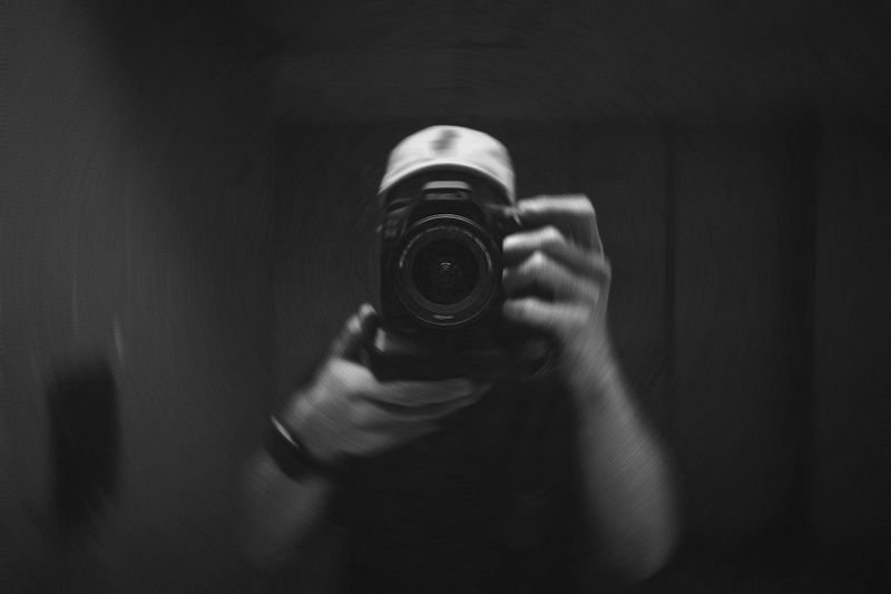 A person in a cap holds a camera, capturing their reflection in a blurred, monochrome image.