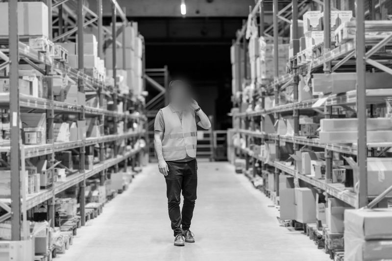 Travailleur portant un gilet de haute visibilité marchant dans une allée d’entrepôt, entouré d’étagères remplies de cartons variés.