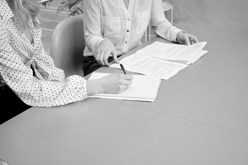 Deux femmes en chemises à pois examinent des documents à une table, l'une écrivant dans un carnet. Image en noir et blanc.
