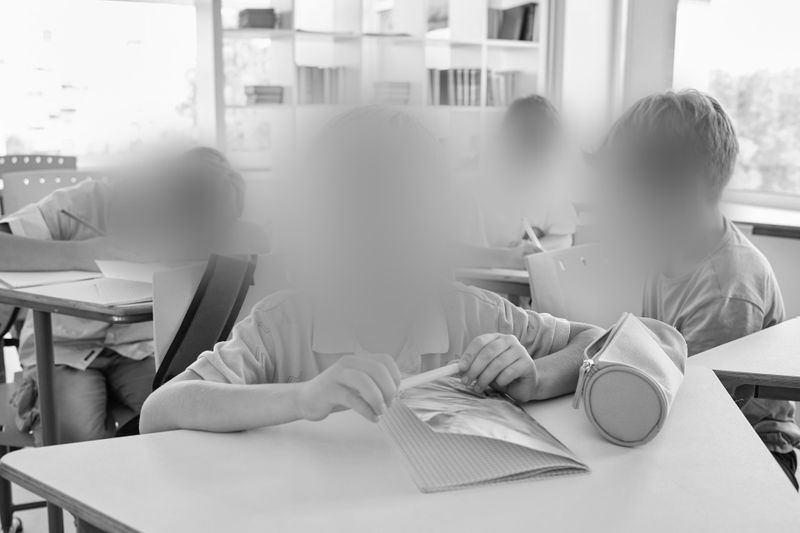 Niños sentados en escritorios en un aula, algunos leyendo libros, con los rostros desenfocados. El entorno es luminoso con luz natural que entra por las ventanas.