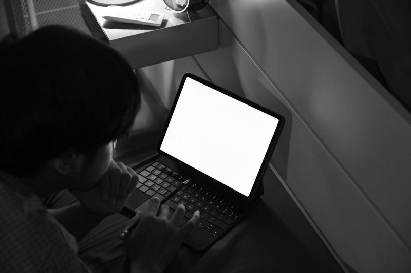 Personne utilisant un ordinateur portable avec un écran lumineux dans une pièce faiblement éclairée, assise sur le sol près d'un bureau.