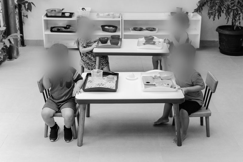 Image en noir et blanc d’enfants assis à de petites tables avec des plateaux, dans une salle de classe avec des étagères et des plantes en arrière-plan.