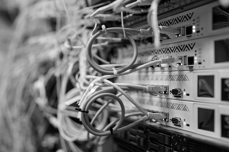 Schwarz-weißes Bild eines Server-Racks mit zahlreichen an Netzwerkausrüstung angeschlossenen Kabeln, die ein komplexes Wirrwarr aus Drähten erzeugen.