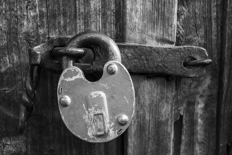 Close-up of a weathered padlock securing a wooden door. The metal is rusted, and the wood appears aged and textured. Black and white image.
