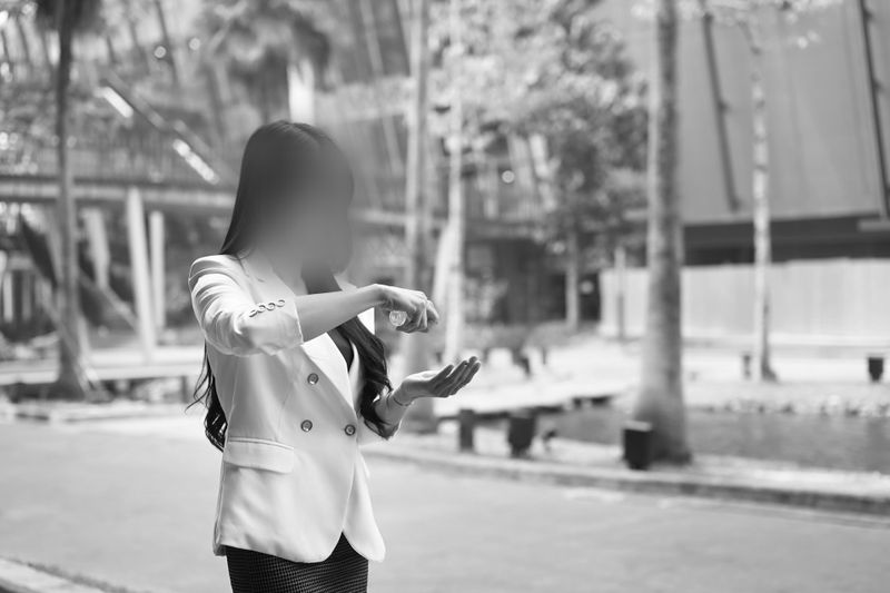 Une femme en blazer blanc se désinfecte les mains à l'extérieur, avec des bâtiments et des arbres flous en arrière-plan. Image en noir et blanc.