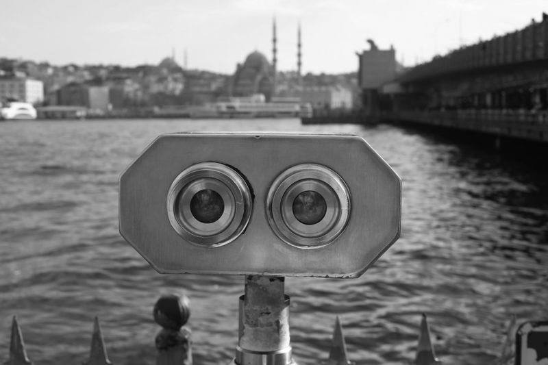 Coin-operated binoculars facing a waterfront with a city skyline and mosques in the background, in black and white.