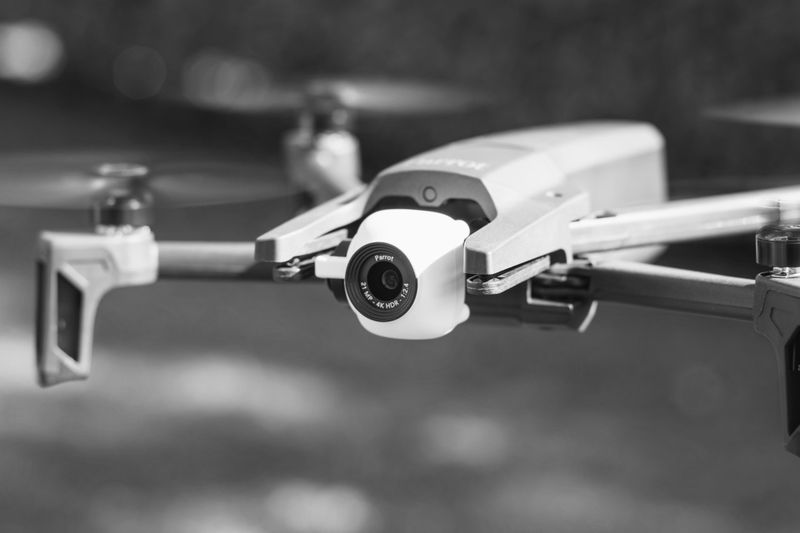 Close-up of a quadcopter drone in flight, showing its camera and propellers, set against a blurred outdoor background in black and white.