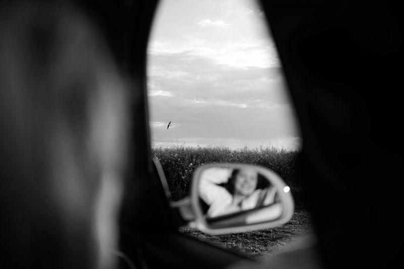 foto en blanco y negro desde el interior del coche, en el espejo se refleja una pasajera y, de fondo, a través de la ventana abierta, el campo y el cielo