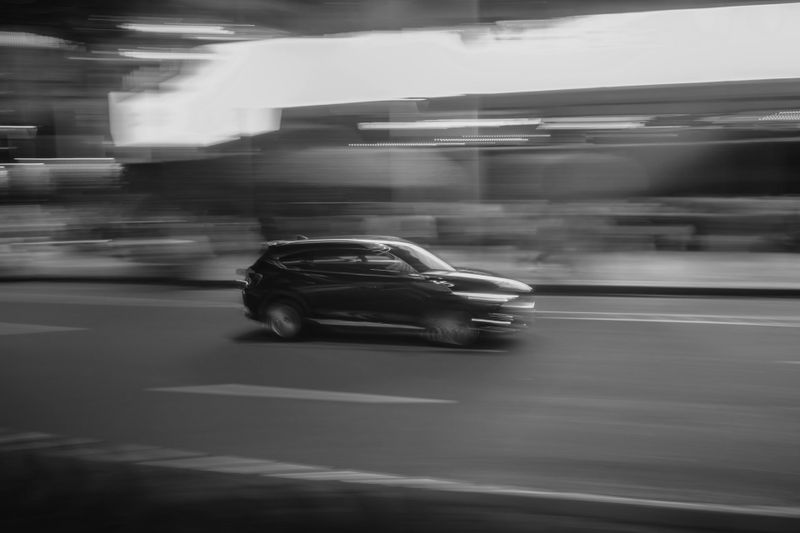 Black and white image of a car speeding down a city street at night, with motion blur emphasizing its fast movement.