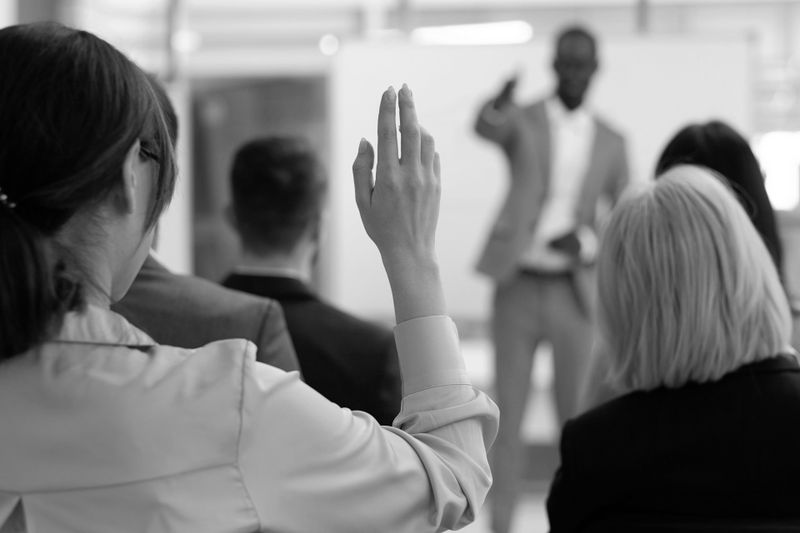 Miembro de la audiencia levantando la mano durante una presentación, con un orador en traje dirigiéndose al grupo. Imagen en blanco y negro.