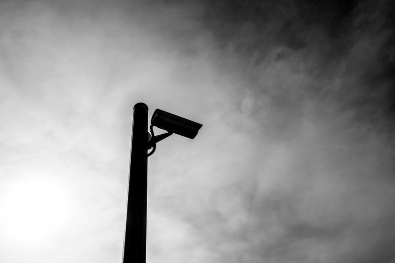 Silhouetted security camera mounted on a pole against a cloudy sky, with the sun partially visible in the background.