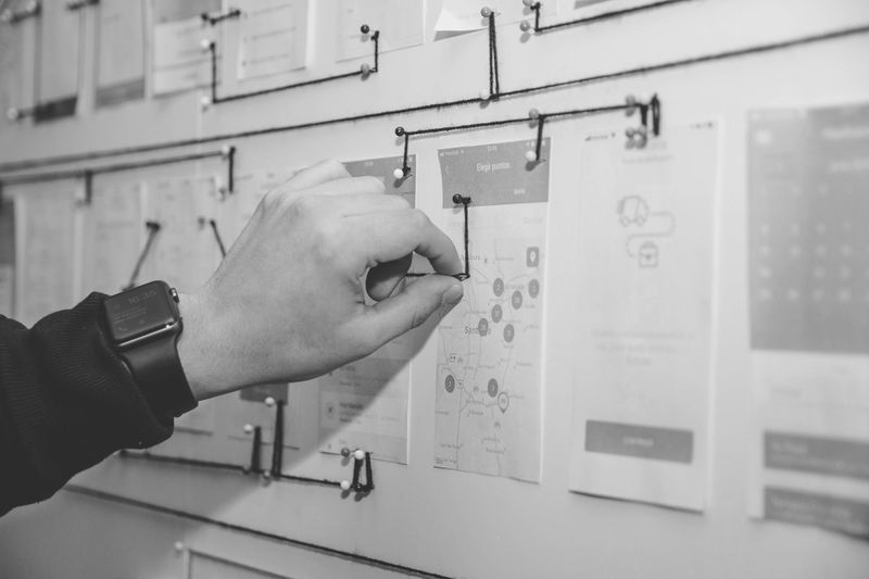 Eine Person mit einer Uhr heftet ein Papier an ein Brett, auf dem mehrere Dokumente mit Fäden verbunden sind, was auf einen Planungsprozess hinweist.