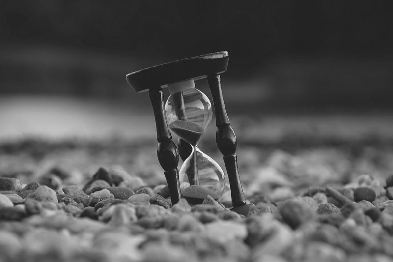 Una imagen en blanco y negro de un reloj de arena descansando sobre un lecho de pequeñas piedras, con arena fluyendo a través de él, sugiriendo el paso del tiempo.