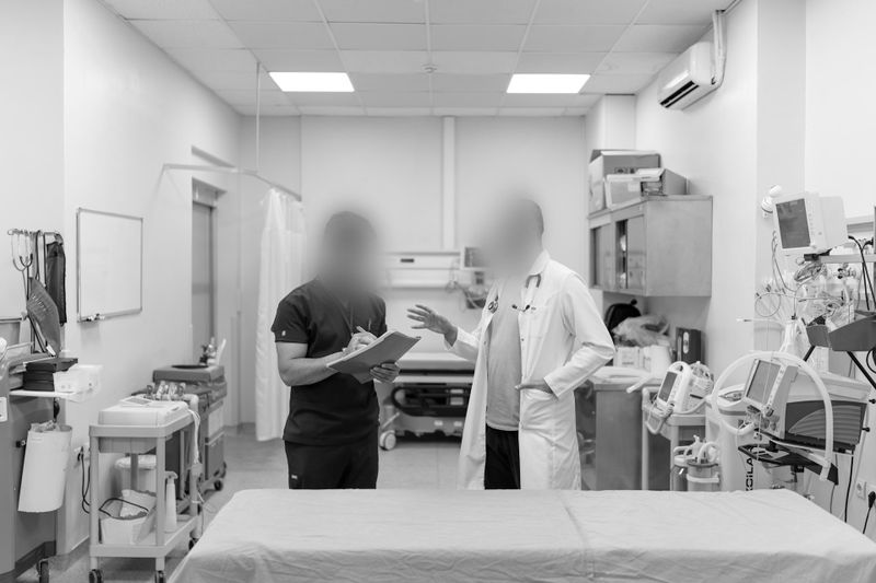 Two medical professionals converse in a hospital room, surrounded by equipment and an examination table, in a grayscale photo.