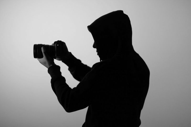 Silhouette d'une personne en sweat à capuche tenant un appareil photo, sur un fond gris uni.