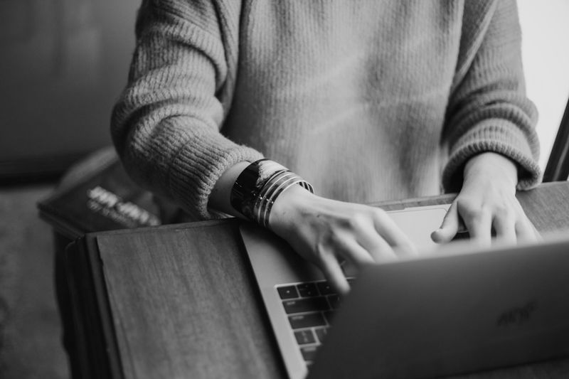Persona con suéter escribiendo en una laptop en un escritorio de madera, con un libro cerca. Imagen en blanco y negro.