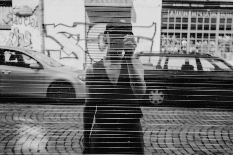 Foto en blanco y negro de una persona tomando una foto a través de una ventana, con coches y grafitis de fondo.