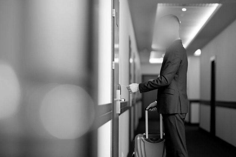 Person in a suit with a suitcase entering a hotel room with a keycard in a hallway, depicted in black and white.