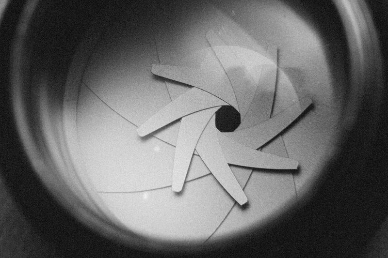 Close-up of a camera lens aperture with overlapping blades forming a circular pattern, creating a geometric design in black and white.