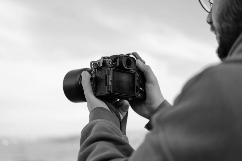 A person holding a DSLR camera and focused on the screen, with a cloudy sky in the background. Black-and-white photo.