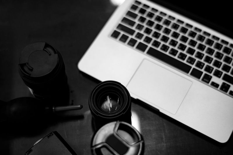 Imagen en blanco y negro de un portátil, lentes de cámara y una herramienta de limpieza de lentes sobre una superficie oscura.