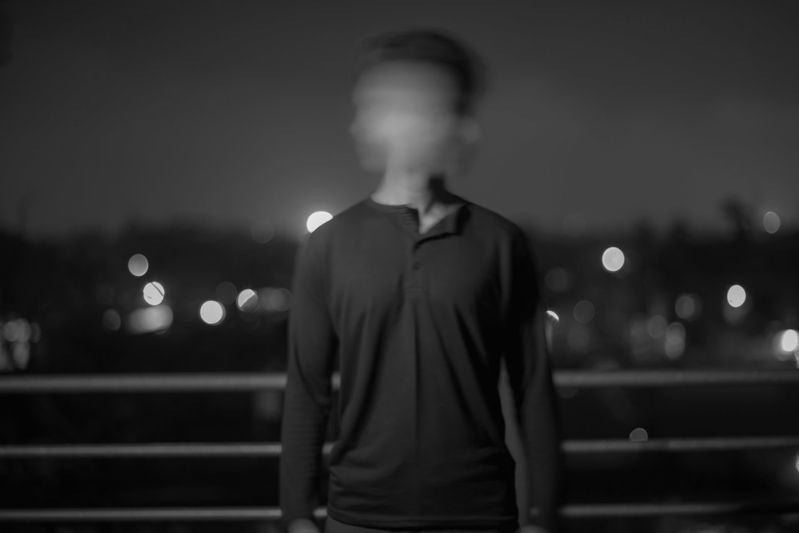 Una persona con el rostro borroso se encuentra frente a un paisaje urbano por la noche, con luces bokeh de fondo. Imagen en blanco y negro.