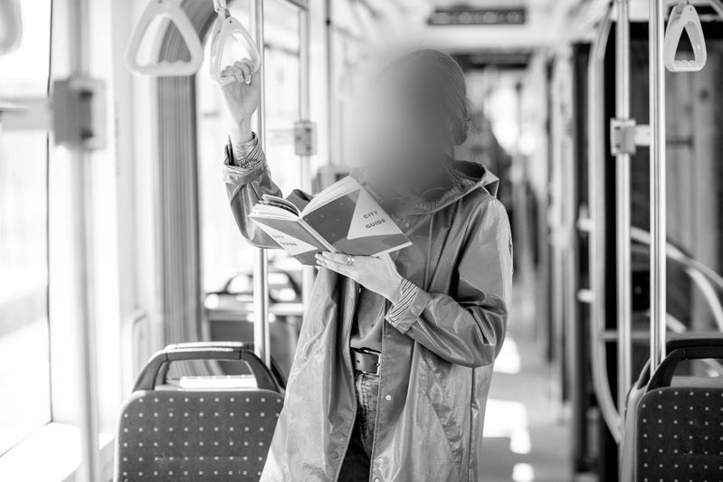Personne en manteau lisant un livre en se tenant debout dans un bus ou un tram, tenant une poignée de soutien. Image en noir et blanc.