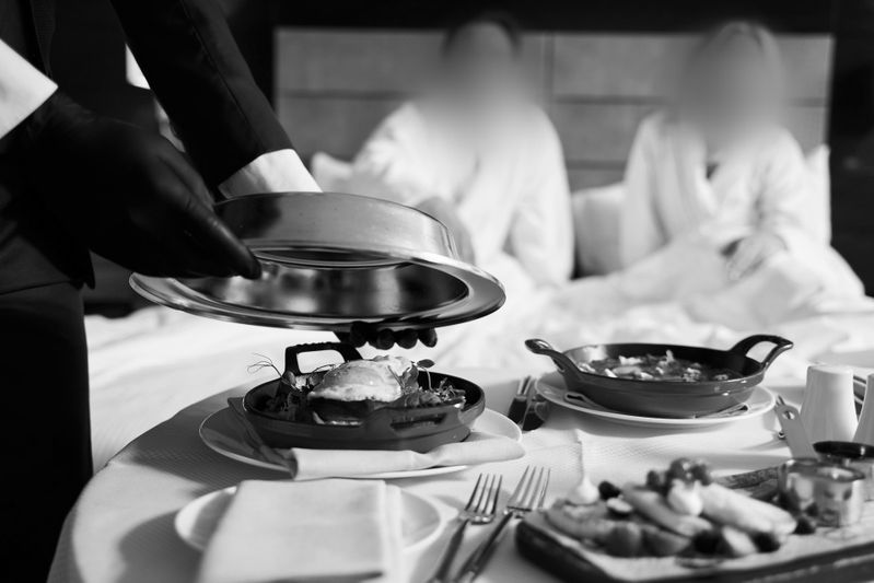 Un serveur, les mains gantées, présente un plat couvert à deux personnes en peignoir assises sur un lit, avec un plateau de petit-déjeuner et des couverts posés sur une table.