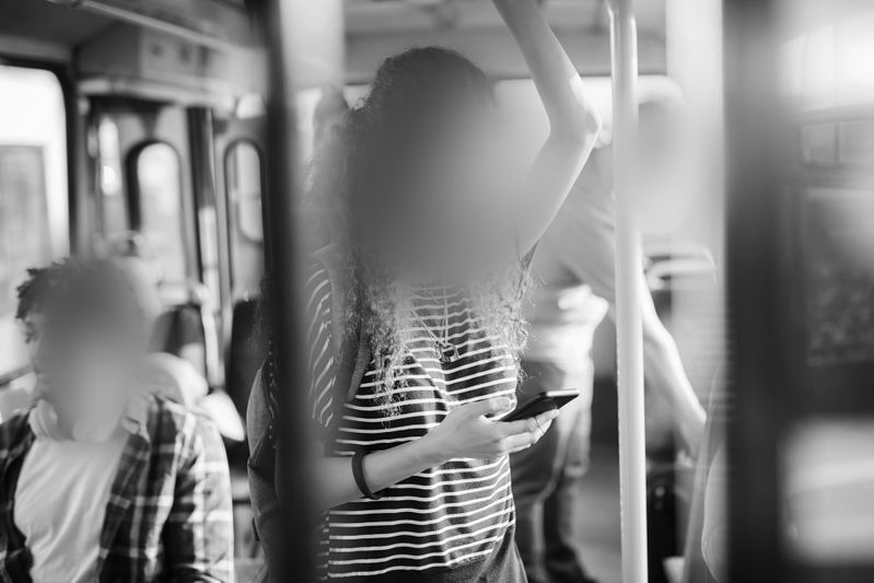 Personne debout dans un bus, tenant un téléphone et s'accrochant à une barre. D'autres passagers sont assis. L'image est en noir et blanc.