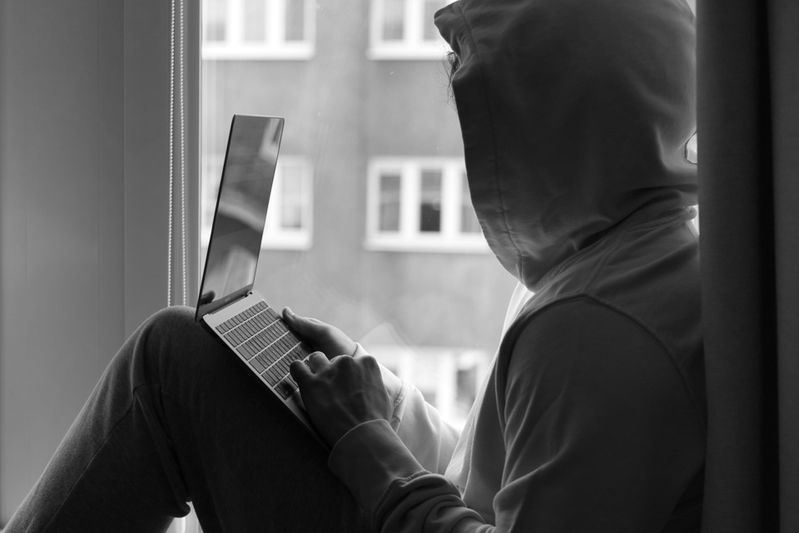 Une personne en sweat à capuche assise près d'une fenêtre utilise un ordinateur portable, avec un bâtiment urbain visible à l'extérieur. Image en noir et blanc.