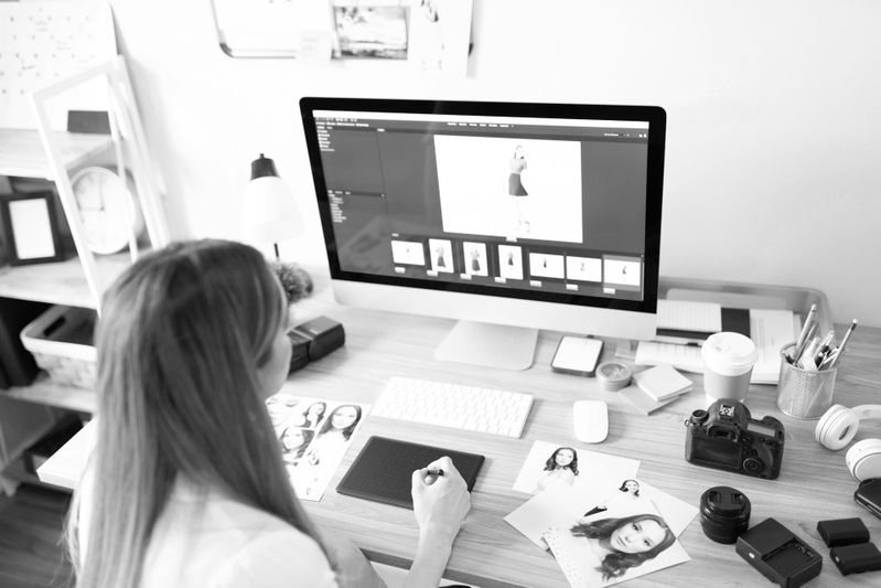 Personne à un bureau utilisant un ordinateur pour la retouche photo, entourée de photographies, d'un appareil photo et d'un café. Noir et blanc.