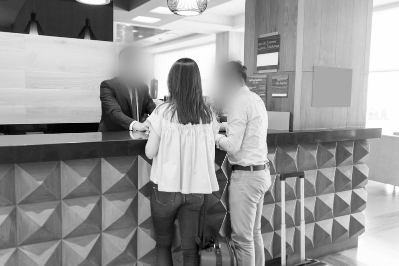 Un couple se tient à la réception d’un hôtel et échange avec un membre du personnel. Des bagages sont posés à côté d’eux. Le cadre est moderne et bien éclairé.