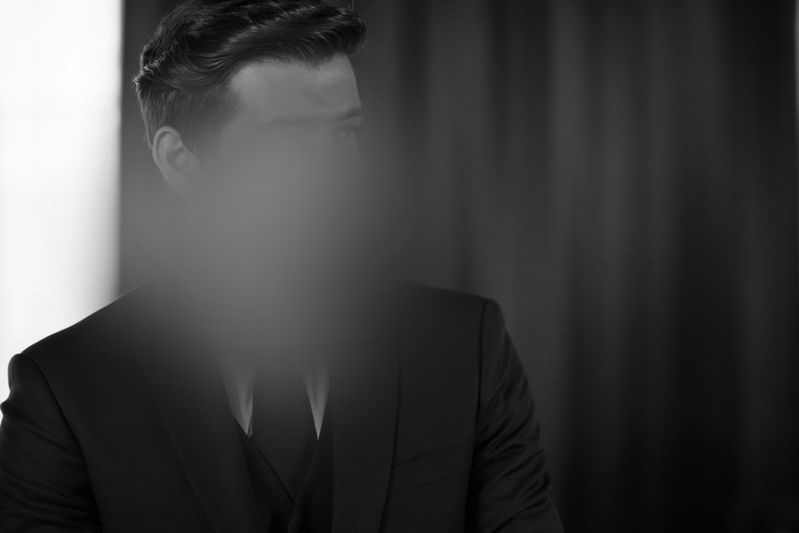 Un hombre con traje y rostro difuso está de pie frente a una cortina oscura; una fotografía en blanco y negro.