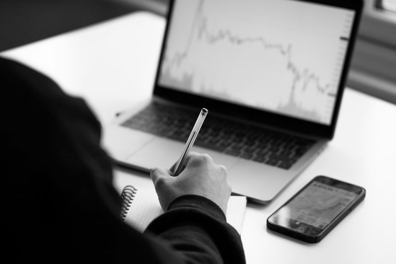 Une personne écrit dans un cahier tout en regardant un ordinateur portable affichant un graphique linéaire. Un smartphone avec un graphique similaire est posé sur la table.