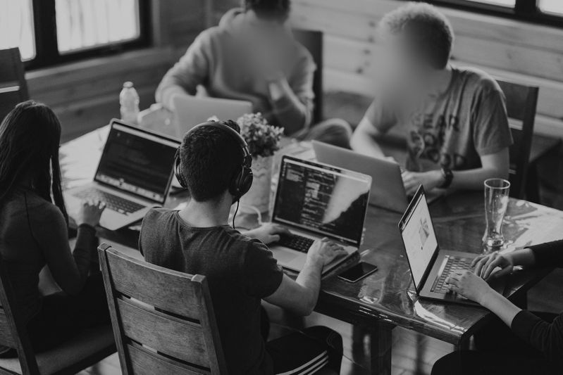 Leute sitzen um einen Tisch und arbeiten an Laptops in einer lockeren Umgebung, einige tragen Kopfhörer.