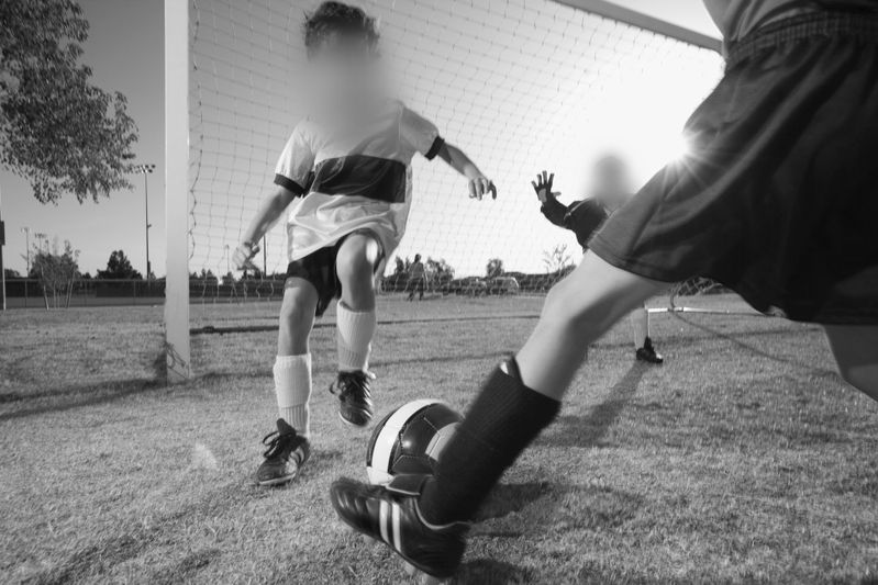 Kids playing soccer, one kicking a ball toward the goal as a goalie moves to block. Sunlight casts long shadows on the grassy field.