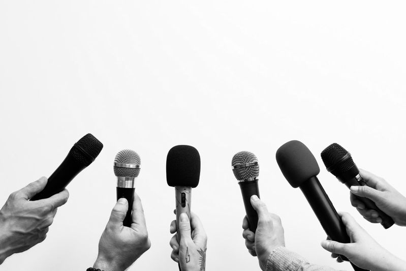 Mains tenant divers microphones sur fond blanc uni, prêtes pour une interview ou une conférence de presse. Image en noir et blanc.