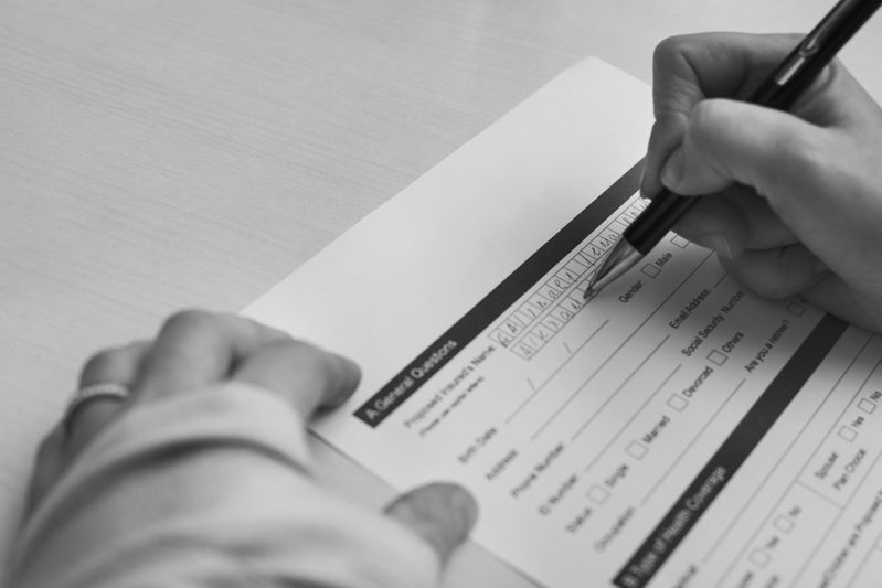 Person filling out a form with a pen, focusing on the signature section. The form is placed on a light-colored table.