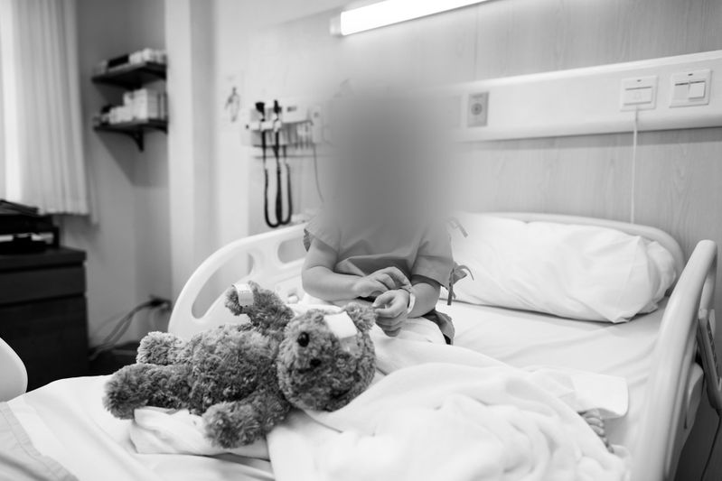 A child with a blurred face sits on a hospital bed, holding an object, with a stuffed animal beside them. The room is equipped with medical devices.