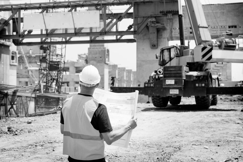 Fotografía en blanco y negro de un trabajador de la construcción con chaleco sobre un fondo de una obra y una máquina de construcción.